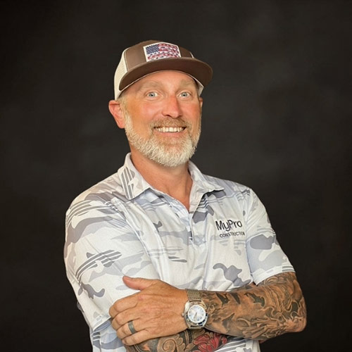 Smiling man with a gray beard wearing a baseball cap and camo-patterned shirt, arms crossed, with visible tattoos and a watch, in front of a dark background.